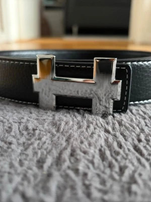 Svart skärp med silverfärgat H-spänne - Snyggt svart bälte i skinn med ett stort, blankt H-format spänne i silver. Bältet har vita sömmar längs kanterna som ger en stilren kontrast. Perfekt accessoar för att lyfta vilken outfit som helst. Helt ny 110cm