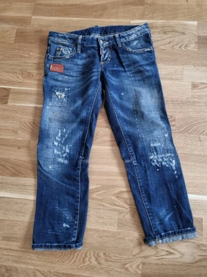 Blå distressed jeans från Dsquared2 -super low  - Säljer ett par blå jeans från Dsquared2 med slitna detaljer och patch på fickan. Super low modell. Jeansen har en klassisk femficksmodell, tydliga slitningar och tvättade partier för en cool, använd look. Materialet är denim i bomull med kontrastsömmar.