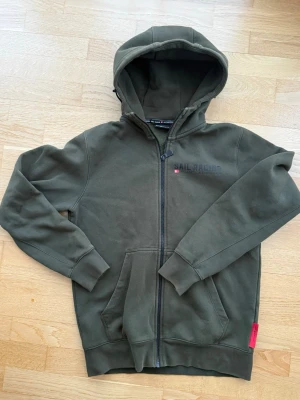 Olivgrön hoodie från Sail Racing - Olivgrön hoodie från Sail Racing med dragkedja framtill och stor huva. Tryck på bröstet med texten 'Sail Racing Antarctica Expedition'. Mjuk insida och känguruficka framtill. Perfekt för en avslappnad och sportig stil.