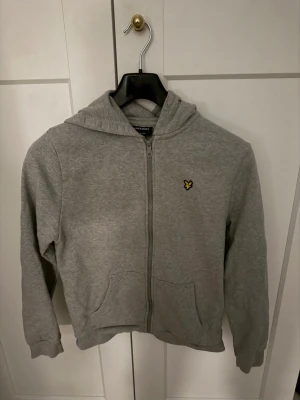 Grå hoodie från Lyle & Scott - Säljer en grå hoodie med dragkedja från Lyle & Scott. Tröjan har huva, två fickor framtill och den klassiska gula loggan på bröstet. Tillverkad i mjukt bomullsmaterial och har ribbade muddar vid ärmar och nederkant. Perfekt för en avslappnad stil.