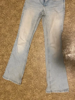 Ljusblå bootcut jeans - Säljer ett par ljusblå bootcut jeans med klassisk femficksdesign och snygga sömmar. Jeansen har utsvängda ben och silverfärgade knappar på bakfickorna. Perfekta för en avslappnad och trendig look.