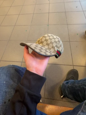 Gucci beige keps med GG-mönster - Säljer en beige keps från Gucci med det klassiska GG-monogrammet över hela kepsen. Kepsen har en böjd skärm och en grön-röd rand på sidan som signaturdetalj. Perfekt för dig som vill ha en exklusiv och trendig accessoar.
