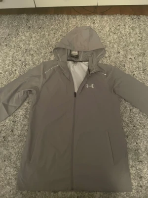 Grå regnjacka från Under Armour - Snygg grå regnjacka från Under Armour med huva och dragkedja framtill. Jackan har två sidofickor och ett diskret logotryck på bröstet. Tillverkad i lätt, vattenavvisande material med meshfoder för extra komfort. Perfekt för regniga dagar. 