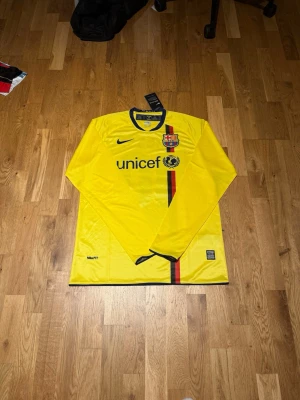 Barcelona 08/09 away kit X Messi #10 - Säljer en gul långärmad FC Barcelona matchtröja från Nike med svart och röd rand på framsidan. Tröjan har klubbmärke, Unicef-logga och är tillverkad i lätt polyester med NikeFIT-teknologi. Perfekt för fotboll och träning.