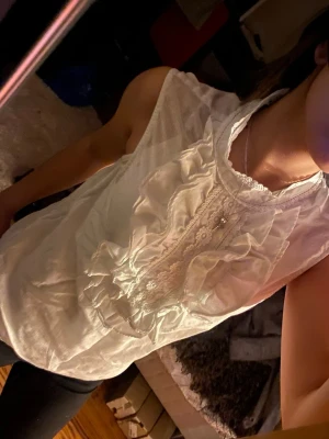 Vit blus med volanger och spets - Söt vit blus med volangdetaljer och spets längs knäppningen framtill. Blusen är ärmlös och har en rundad halsringning med dekorativ kant. Perfekt för dig som gillar romantisk och feminin stil. Materialet känns lätt och luftigt.