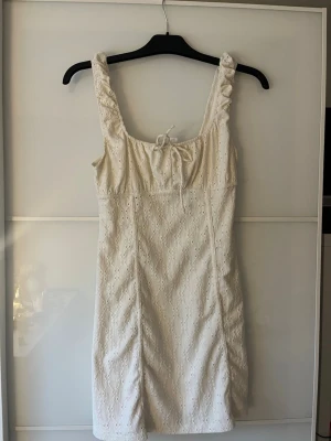 Vit klänning - Söt vit kortklänning med breda och puffiga axelband och dekorativ knytning framtill. Klänningen har ett broderat hålmönster över hela tyget och är tillverkad i ett lätt bomullsmaterial. Perfekt för skolavslutning eller student.