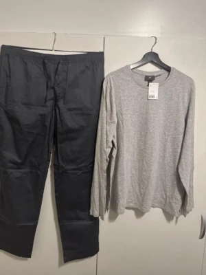 Grå pyjamas från H&M, set med lång ärm - Mjuk och bekväm pyjamas från H&M i set med långärmad ljusgrå tröja och mörkgrå byxor. Tröjan har rund halsringning och byxorna har resår i midjan. Perfekt för avslappnade kvällar och mysiga morgnar.