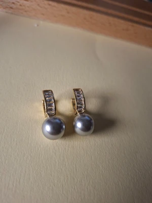 Guldörhängen med pärlor och stenar - Snygga örhängen i guldton med stora silvriga pärlor och rektangulära klara stenar på bygeln. Perfekt för dig som gillar klassisk men ändå trendig stil. Materialet ser ut att vara metall med pärlimitation och glittrande stenar.