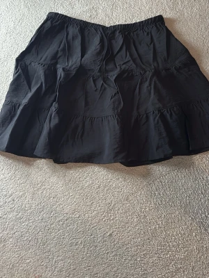 Svart volangkjol med knytband - Säljer en svart kortkjol med volang och knytband i midjan. Kjolen har en avslappnad passform och är gjord i ett mjukt bomullstyg. Perfekt för dig som gillar en enkel men snygg stil. Hör av dig vid frågor!🥰✨