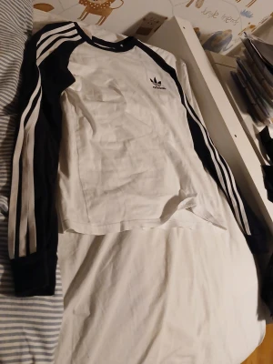 Vit långärmad Adidas t-shirt - Säljer en vit långärmad t-shirt från Adidas med svarta ärmar och klassiska tre ränder längs ärmarna. Adidas-loggan är broderad på bröstet. Tröjan har en sportig look och är gjord i mjuk bomull för maximal komfort. Priset kan diskuteras.
