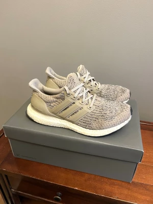 Adidas Ultraboost Beige - Varsamt använda Adidas Ultraboost sneakers i storlek 39, beige och grå toner med stickad ovandel och vita Boost-sulor. Skorna har klassiska tre ränder på sidan och matchande beige snörning. Perfekta för dig som gillar stilrena och bekväma sneakers med modern design. Säljer billigt för har ingen plats för dem.