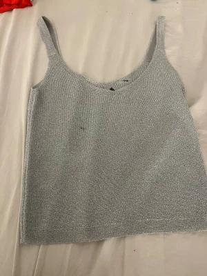 Silver stickad linnetopp - Snygg silverfärgad stickad linnetopp med breda axelband och rundad halsringning. Perfekt för att skapa en cool och glittrig look. Materialet är stickat och ger en skön känsla mot huden. Passar dig som vill ha något enkelt men ändå lite extra.