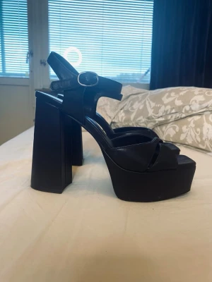 Svarta platåpumps med blockklack - Säljer ett par svarta platåpumps med blockklack och öppen tå. Skorna har en rem över foten och justerbart spänne vid ankeln. Tillverkade i glansigt material som ger en cool och trendig look.