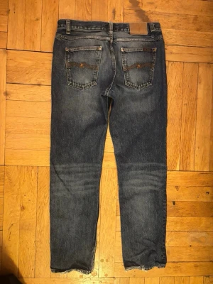 Mörkblå jeans från Nudie Jeans - W32 L32 Säljer ett par mörkblå jeans från Nudie Jeans med klassisk femficksdesign och raka ben. Jeansen har snygga slitningar och kontrastsömmar samt en patch bak i midjan. Tillverkade i slitstark denim, perfekta för en avslappnad stil.