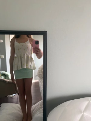 Vit blommig linnetopp med volang - Supersöt vit linnetopp med gula blommor och volangkant. Toppen har smock upptill, tunna axelband och ett flowy fall. Perfekt för varma dagar och ger en fräsch sommarkänsla. Materialet känns lätt och luftigt.