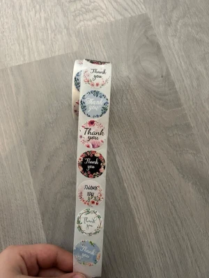 Rulle med Thank you-klistermärken - En rulle med runda klistermärken i olika färger och blommiga mönster, alla med texten 'Thank you'. Perfekt för paket, brev eller presenter. Klistermärkena har olika bakgrunder som svart, blå, rosa och vit med fina blomdetaljer.