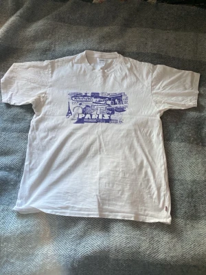 Vit t-shirt från Levis  - Säljer en vit t-shirt med ett stort blått tryck på framsidan. Den är från Levis och är i storleken S men sitter som en M! Jätte bra skick och inte använd alls många gånger.