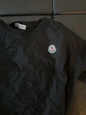 Svart Moncler t-shirt - Snygg svart t-shirt från Moncler med klassisk rund hals och det ikoniska Moncler-märket broderat på bröstet. Enkel och stilren design som passar till allt. Tillverkad i mjuk bomull för skön känsla.