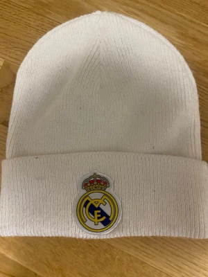 Vit Adidas Real Madrid mössa - Säljer en vit ribbstickad mössa från Adidas med Real Madrid-logga framtill och Adidas-logga i guld på baksidan. Klassisk beanie-modell med uppvikt kant, perfekt för dig som vill visa ditt stöd för Real Madrid även under kalla dagar.