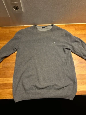 Grå stickad tröja från Gant - Snygg grå stickad tröja från Gant med rund halsringning och diskret logga broderad på bröstet. Tröjan har långa ärmar och ribbade muddar vid ärmslut och nederkant. Perfekt för lager på lager och enkel att matcha med olika outfits. 