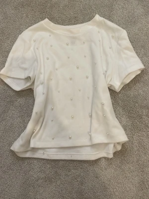 Vit topp med pärlor från H&M - Säljer en vit topp från H&M med korta ärmar och dekorativa pärlor över hela framsidan. Toppens material är mjukt och bekvämt, perfekt för att ge en snygg touch till din outfit. Enkel rund halsringning och något loose passform. Storlek S men eftersom Divided (H&M:s tonårsmärke) är för tonåringar passar tröjan som xxs/xs i vanlig vuxenstorlek❤️