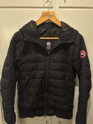 Canada Goose cardigan  - Snygg svart cardigan från Canada Goose med huva och dragkedja framtill. Jackan har quiltad design, ribbade muddar och den klassiska logotypen på ärmen. Perfekt till våren. Priset kan såklart alltid diskuteras. Hör av dig vid frågor eller funderingar.