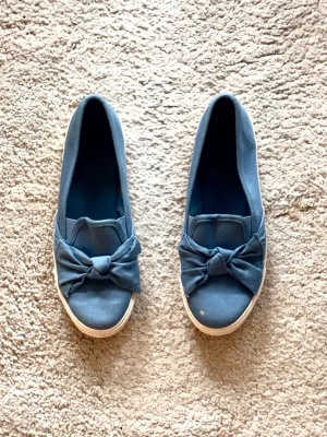 Denim ballerinaskor med rosett  - Säljer ett par snygga blå ballerinaskor i jeanstyg/ denim med en stor rosett framtill. Skorna har vit sula och slip-on-modell som gör dem enkla att ta på. Perfekta för dig som gillar en söt och avslappnad stil.