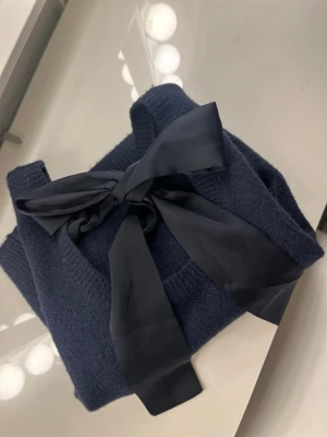 Mörkblå stickad topp med rosett från Neo Noir - Snygg mörkblå stickad topp med korta puffärmar och en stor svart rosett framtill vid halsen. Materialet är mjukt och ger en skön känsla. Perfekt för dig som gillar detaljer och vill ha något unikt i garderoben.