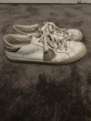 Vita sneakers med glitterdetaljer - Säljer ett par vita låga sneakers med silverfärgade glitterdetaljer på hälen och grå stjärna på sidan från Philippe modell. Skorna har klassisk snörning och platt sula. Perfekta för dig som gillar en touch av bling i vardagen. Ens är rengjord men ska göra rent dom ordentligt innan dom skickas såklart!