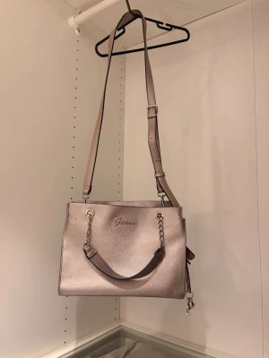 Guess metalliclila handväska - Handväska från Guess i metallicrosa med både axelrem och handtag. Väskan har flera fack, dragkedja och magnetlås. Perfekt för dig som vill ha en stilren och trendig accessoar. Man får plats med mycket tex stor parfym, hårborste. Lite slitage på överkanten.