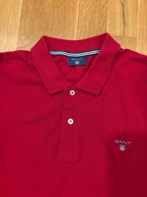 Röd långärmad polotopp från GANT - Snygg röd långärmad polotopp från GANT med klassisk krage och två knappar framtill. Toppens material är bomull och den har logotyp broderad på bröstet. Perfekt för dig som gillar stilrena och tidlösa plagg.