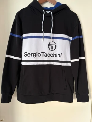 Svart hoodie från Sergio Tacchini - Cool svart hoodie med vit och blåa detaljer från Sergio Tacchini. Stor logga och text framtill, huva med vita snören och ribbade muddar. Materialet är mjukt och bekvämt, perfekt för en avslappnad stil.