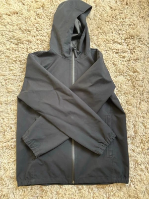 Svart regnjacka från Uniqlo - Snygg svart regnjacka från Uniqlo med huva och hel dragkedja framtill. Jackan har två sidofickor och elastiska muddar vid ärmsluten. Tillverkad i ett vattenavvisande material som passar perfekt för regniga dagar. Passar 158-165cm