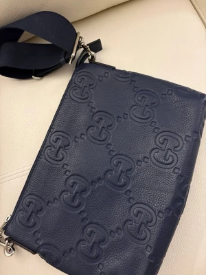 Mörkblå axelväska från Gucci - Snygg mörkblå axelväska från Gucci i präglat skinn med GG-logga över hela väskan. Väskan har bred justerbar axelrem i textil och dragkedja med metall-detaljer. Perfekt för dig som vill ha en stilren och lyxig accessoar.