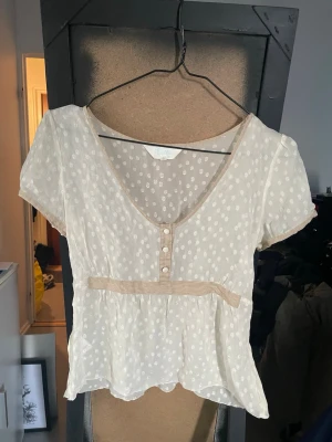 Prickig blus från Evita med knappar - Söt, ljusbeige blus från Evita med vita prickar, korta ärmar och dekorativa knappar framtill. Blusen har en markerad midja med ett band och lätt v-ringad hals. Tillverkad i ett tunt, transparent material som ger en luftig känsla.