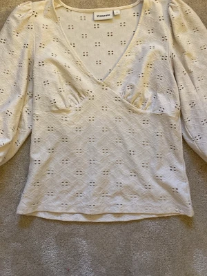Vit broderad blus från Kappahl - Säljer en vit blus från Kappahl med omlott-design och v-ringning. Blusen har lite längre ärmar och är tillverkad i bomull med broderade detaljer och små hålmönster. Perfekt för dig som gillar romantiska och luftiga toppar.