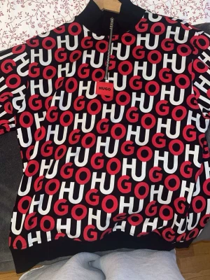 Svart och röd Hugo Boss hoodie - Säljer en svart hoodie från Hugo Boss med halv dragkedja och ett fett all-over tryck i rött och vitt där det står HUGO. Ribbad mudd vid ärmslut och nederkant. Materialet är mjukt och bekvämt, perfekt för en streetwear-look.