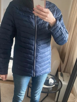 Mörkblå jacka med dragkedja storlek S-M - Mörkblå dunjacka med quiltad design och hög krage. Jackan har hel dragkedja framtill och raka ärmar, vilket ger en clean och modern look. Perfekt för kalla vinterdagar och enkel att styla till olika outfits.