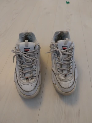 Vita sneakers från Fila - Klassiska vita sneakers från Fila med chunky sula och logga på plös och häl. Skorna har vita snören och detaljer i rött och blått på loggan. Perfekt för en sportig och trendig look. Lite slitet i tyget bak. Helt ok skick. Se bilder. Använda av en tjej. Ganska små I storleken.