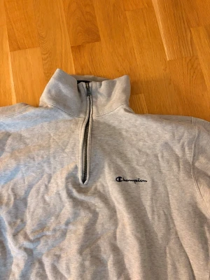 Grå halvzip sweatshirt från Champion - Snygg grå sweatshirt från Champion med halv dragkedja och hög krage. Tröjan har broderad logga på bröstet och ribbade muddar vid ärmslut och nederkant. Perfekt för en avslappnad och sportig stil.