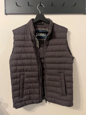 Brun dunväst från Herno - Snygg brun dunväst från Herno med quiltad design och hög krage. Västen har dragkedja framtill och två sidofickor med knappar. Perfekt för lager-på-lager och ger en clean look till din outfit. Tillverkad i lätt och värmande material. Den är dessutom i nyskick och är endast använd några fåtal gånger.