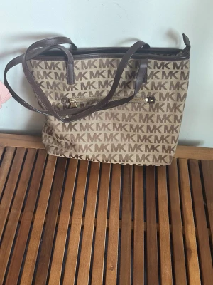 Brun MK handväska med logomönster - Snygg handväska från Michael Kors i beige och brunt med MK-logga över hela väskan. Väskan har bruna handtag, guldfärgad dragkedja och en praktisk ytterficka. Perfekt för dig som gillar klassisk design och vill ha plats för allt du behöver.