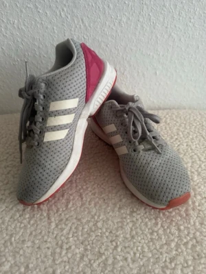 Grå Adidas sneakers med rosa detalj - Snygga grå sneakers från Adidas med klassiska vita ränder på sidorna och rosa detalj vid hälen. Ovandelen är i meshmaterial för bra andningsförmåga och sulan har inslag av vitt och rosa. Perfekta för en sportig och avslappnad stil.