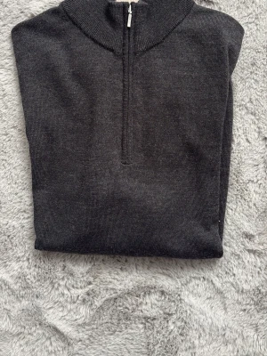 Mörkgrå stickad halvzip-tröja - Snygg mörkgrå stickad tröja med halv dragkedja och ribbad krage. Perfekt till lager på lager och enkel att matcha med olika outfits. Tröjan har långa ärmar och ribbade muddar vid ärmslut och nederkant.