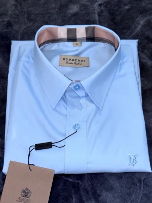Ljusblå skjorta från Burberry - Klassisk ljusblå skjorta från Burberry med ikonisk rutdetalj på insidan av kragen och diskret broderad logga vid nederkanten. Skjortan har knappar framtill och är tillverkad i mjuk bomull för en clean och stilren look.