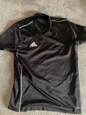 Svart Adidas sporttröja herr - Svart sporttröja från Adidas med vita detaljer och logga på bröstet. Tillverkad i lätt och snabbtorkande material, perfekt för träning eller match. Klassisk passform med korta ärmar och rund hals. Aeroready-teknologi för extra komfort.