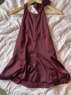 Vinröd halterneckklänning med rosett - Säljer en vinröd kortklänning i halterneckmodell med stor rosettdetalj i nacken. Klänningen har en lätt glansig finish och är ärmlös med öppen rygg. Perfekt för dig som vill ha en elegant och trendig look.