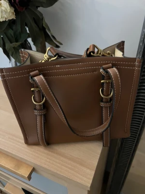 Brun fyrkantig handväska i läderlook - Snygg brun handväska i läderimitation med vita kontrastsömmar och guldfärgade metalldetaljer. Väskan har både handtag och en justerbar axelrem, samt en stilren, fyrkantig form. Perfekt för dig som gillar klassisk men trendig design.