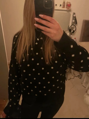 Svart stickad tröja med vita prickar - Snygg svart stickad tröja med vita prickar över hela plagget. Tröjan har en avslappnad passform, rund halsringning och långa ärmar. Perfekt för dig som gillar en trendig och mysig look. Materialet är mjukt, inte alls stickigt.