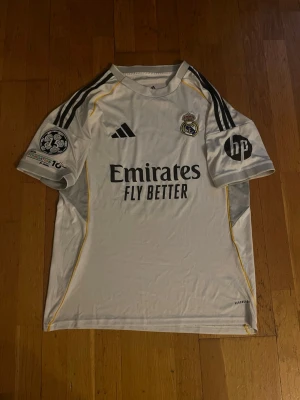 Real Madrid matchtröja Bellingham - Vit Real Madrid matchtröja med svarta detaljer och gula inslag. Adidas-logga, klubbmärke och sponsortryck på framsidan. Nummer 5 och Bellingham på ryggen. Korta ärmar och rund hals. Perfekt för fotbollsfans! Använd 3-4 gånger 